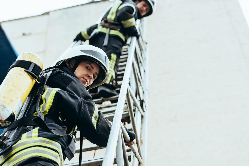 nízký úhel pohledu hasičů v přilbách, stojící na žebříku a při pohledu na fotoaparát - Fotografie, Obrázek
