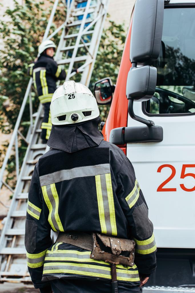 Achteraanzicht van brandweerman in beschermende uniform en helm collega kijken op de ladder op straat - Foto, afbeelding