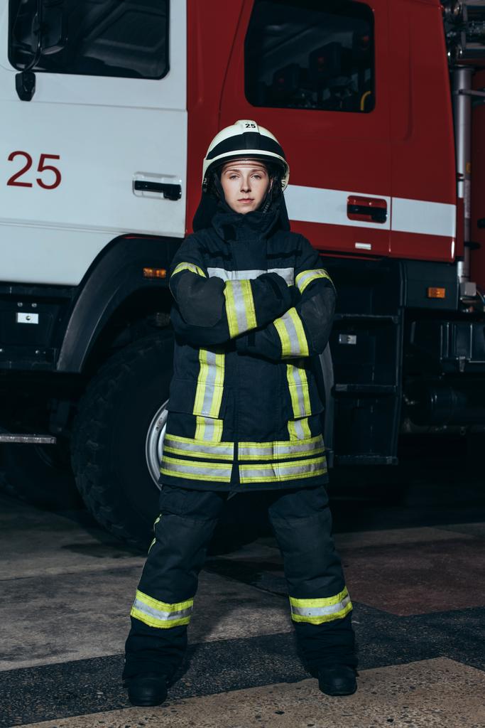 Feuerwehrfrau im Helm mit gekreuzten Armen steht am Feuerwehrhaus mit LKW dahinter - Foto, Bild