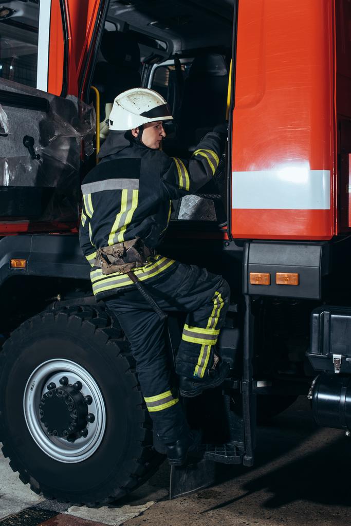 brandweerman in beschermende uniform en helm krijgen in vrachtwagen bij brandweerkazerne - Foto, afbeelding
