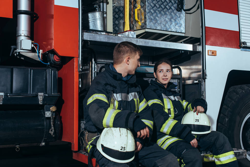 Brandweerlieden in beschermend uniform in gesprek nabij vrachtwagen bij brandweerkazerne - Foto, afbeelding