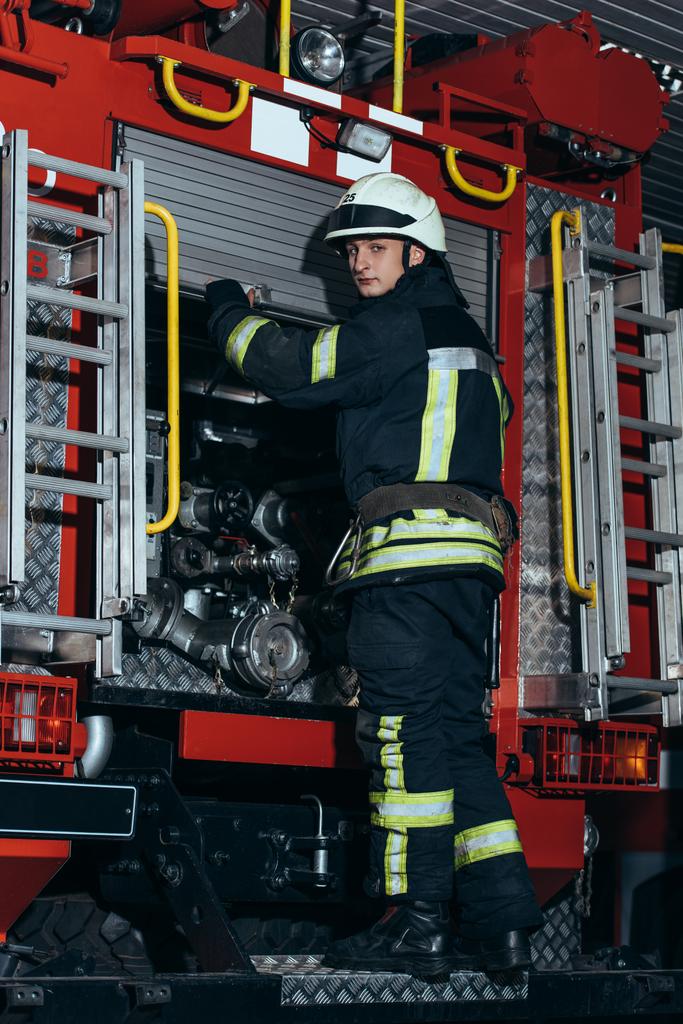 Feuerwehrmann in Schutzuniform und Helm blickt in Feuerwehrhaus auf Kamera - Foto, Bild