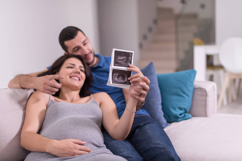 junges schwangeres Paar sucht Baby-Ultraschall-Foto, während es sich zu Hause auf dem Sofa entspannt - Foto, Bild