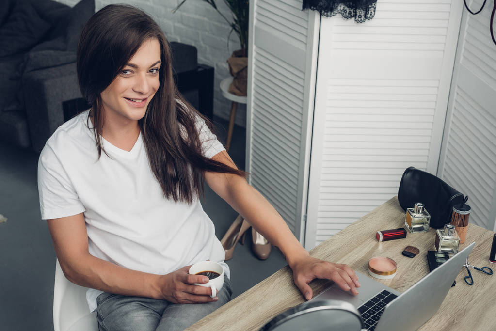 uśmiechnięta młoda transgenderowa freelancerka z filiżanką kawy siedząca w domu w miejscu pracy i patrząca w kamerę - Zdjęcie, obraz