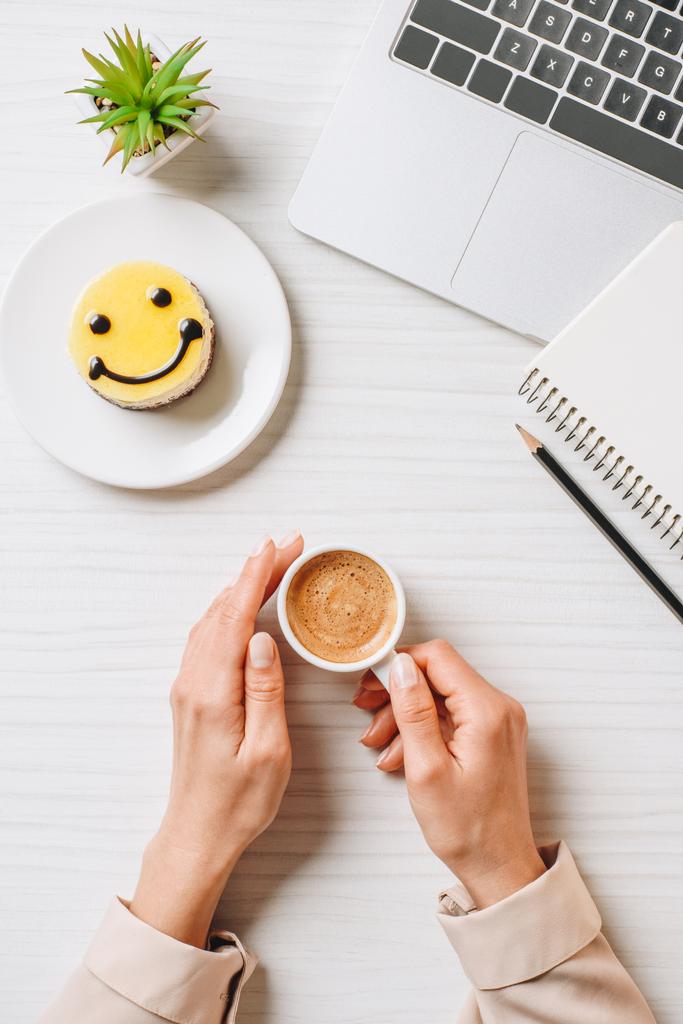 gedeeltelijke weergave van zakenvrouw drinken koffiekopje aan tafel met laptop en cake met symbool van glimlach in office - Foto, afbeelding
