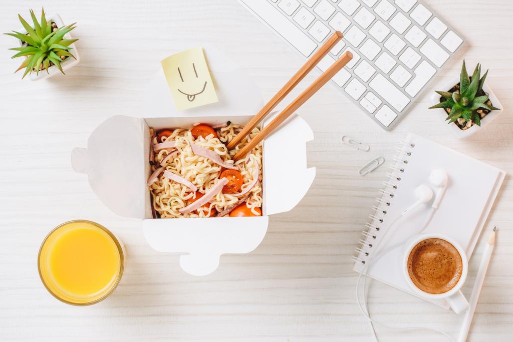 elevated view of wok box with chopsticks, orange juice, symbol of smile and coffee cup at table in office  - Photo, Image