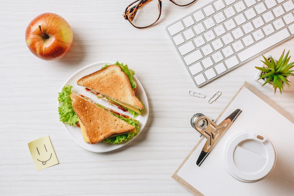 вид зверху на робоче місце з бутербродом, паперовою чашкою кави, яблуком і символом посмішки за столом в офісі
  - Фото, зображення