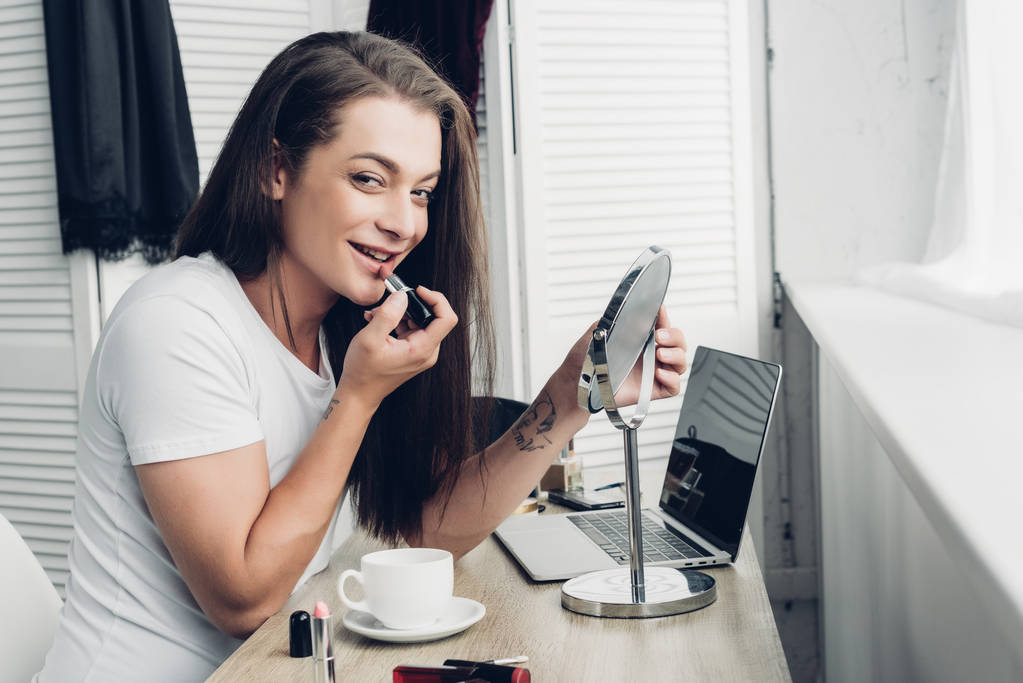 smiling young transgender woman with round makeup mirror applying lipstick while looking at camera - Photo, Image
