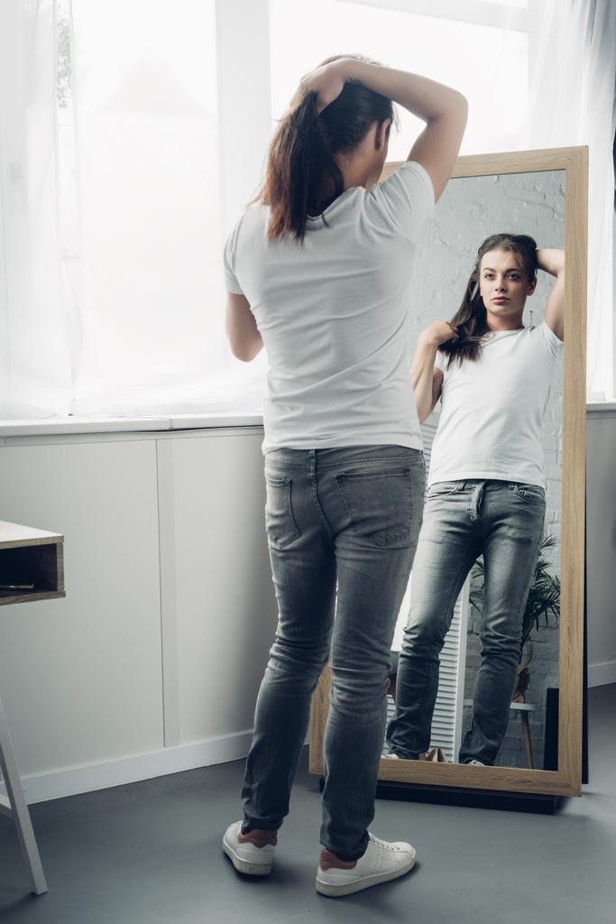 трансгендерная женщина в белой футболке смотрит в зеркало дома - Фото, изображение