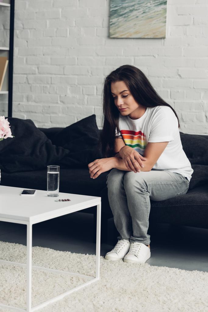 junge Transgender-Frau sitzt zu Hause auf Couch und schaut sich Tabletten und Wasser an - Foto, Bild