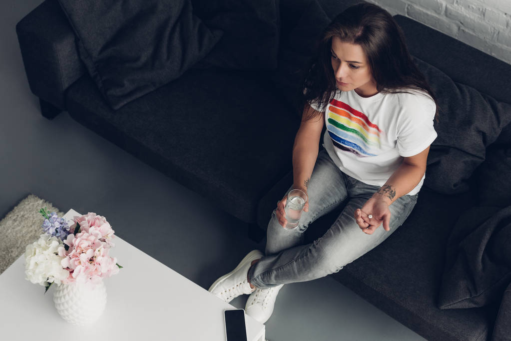 высокий угол обзора молодой трансгендерной женщины с таблетками и стаканом воды, сидящей на диване - Фото, изображение