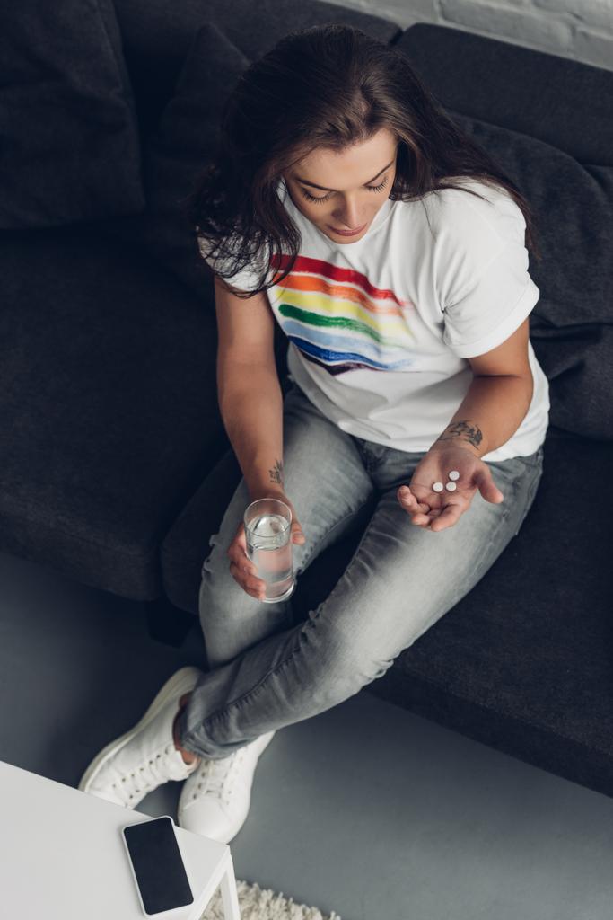 うつ病の若いトランスジェンダーの女性の角度の高さと錠剤とコップの水がソファに座っています - 写真・画像
