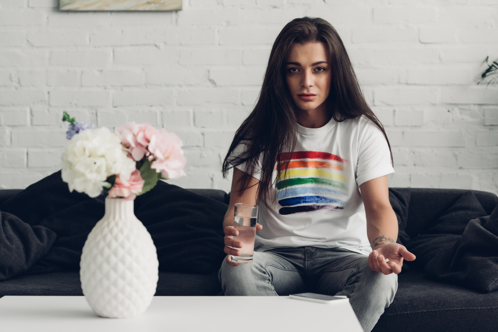 депрессивная трансгендерная женщина с таблетками и стаканом воды, сидящая на диване - Фото, изображение