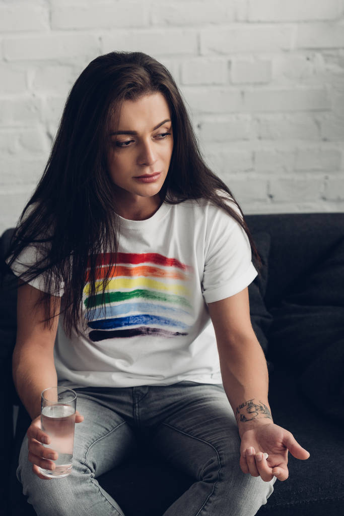 депрессивная молодая трансгендерная женщина с таблетками и стаканом воды, сидящая на диване - Фото, изображение