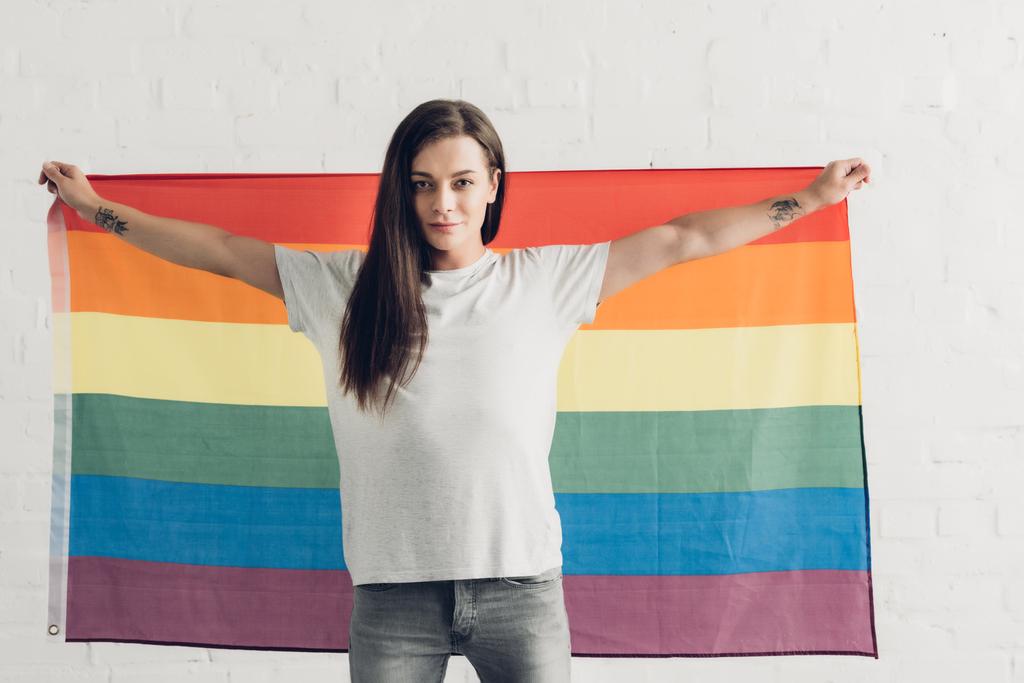 白いレンガの壁の前にプライド旗を掲げる自信に満ちたトランスジェンダーの女性 - 写真・画像