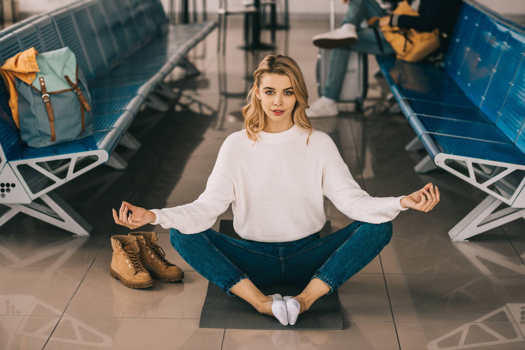 Mädchen meditiert in Lotusposition und blickt in die Kamera, während sie im Flughafenterminal auf ihren Flug wartet  - Foto, Bild