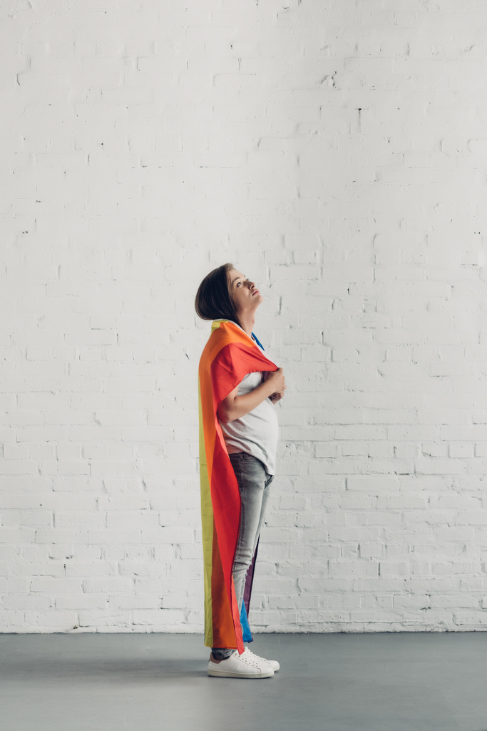 Junge Transgender-Frau bedeckt Schultern mit Stolz-Fahne vor weißer Backsteinwand - Foto, Bild