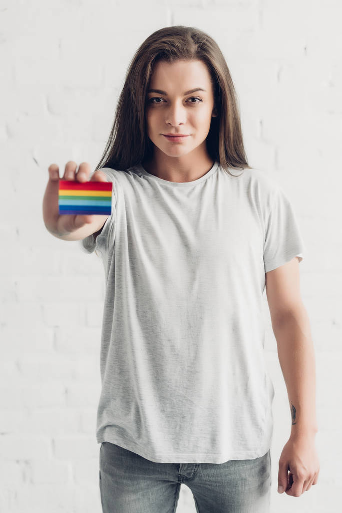 jong transgender vrouw holding card met trots vlag in de voorkant van witte baksteen muur - Foto, afbeelding