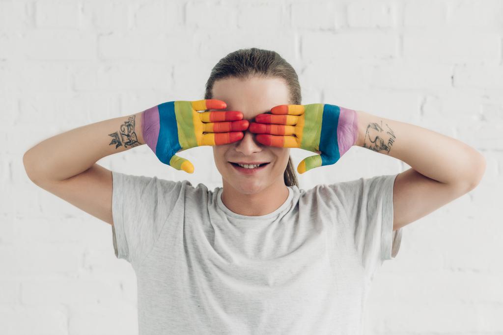 mujer transexual sonriente cubriendo los ojos con las manos pintadas en colores de la bandera del orgullo delante de la pared de ladrillo blanco - Foto, Imagen