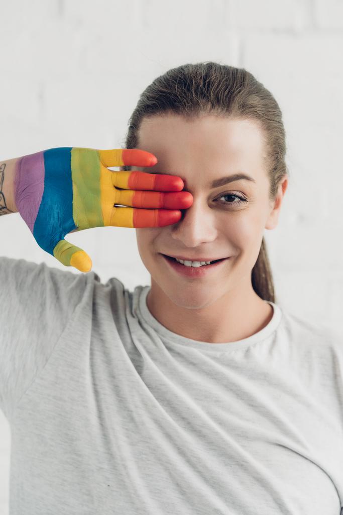 souriant transgenre femme couvrant oeil avec la main peint dans des couleurs de drapeau de fierté en face du mur de briques blanches - Photo, image