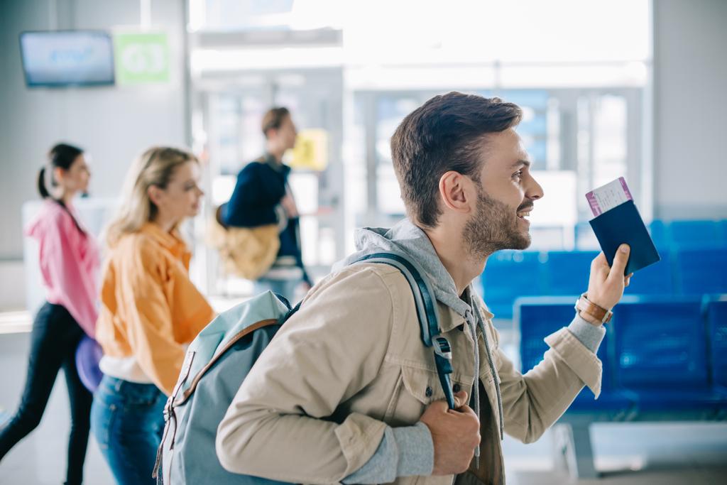 vue latérale de l'homme souriant avec sac à dos tenant passeport et carte d'embarquement à l'aéroport
 - Photo, image