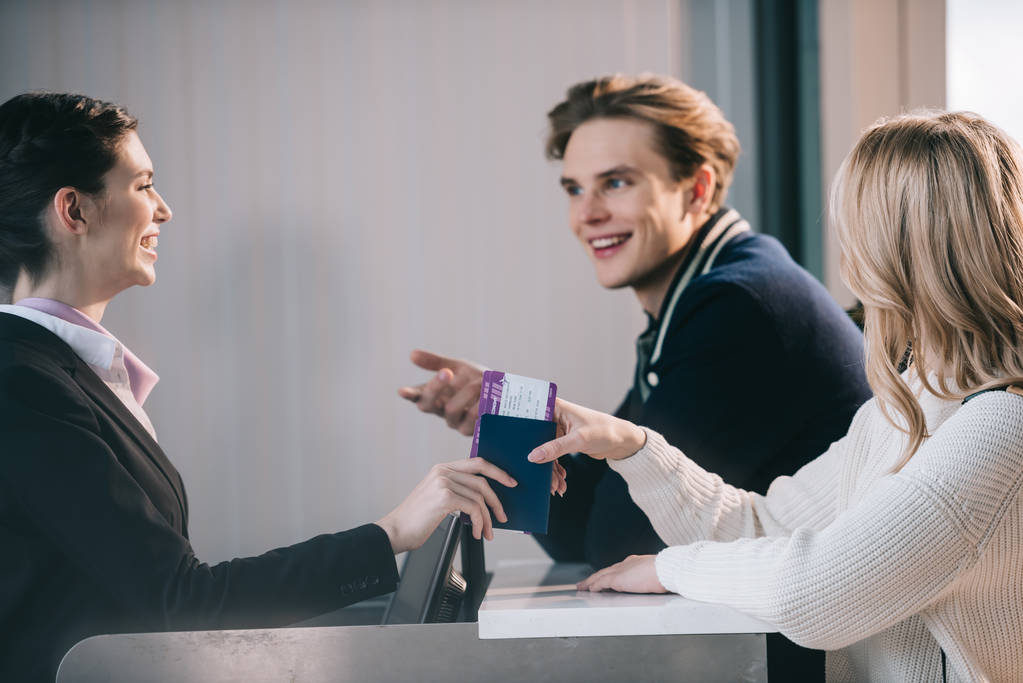 улыбающаяся молодая пара смотрит на работника за стойкой регистрации в аэропорту
 - Фото, изображение
