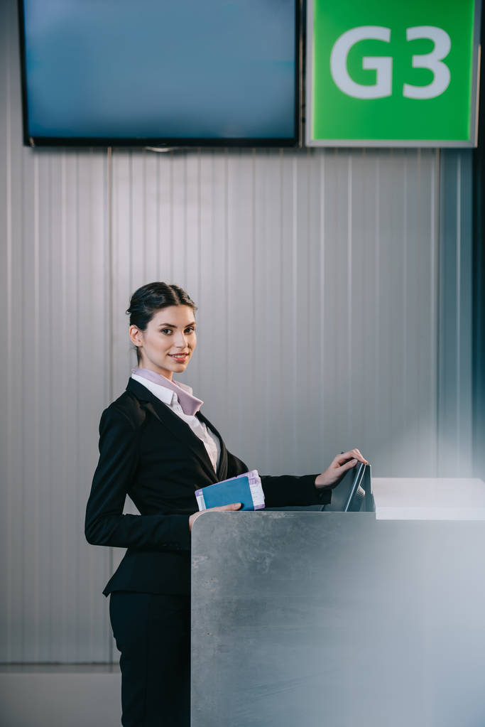 hermosa joven trabajador sosteniendo documentos y sonriendo a la cámara en el mostrador de facturación en el aeropuerto
 - Foto, Imagen