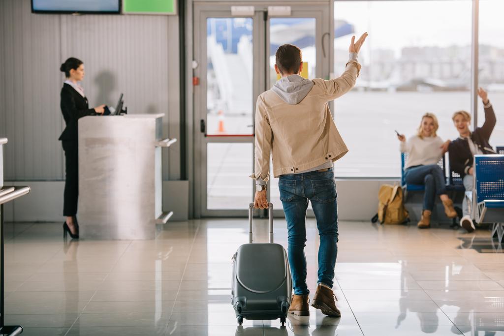 vista trasera del joven con la maleta saludando a los amigos en el aeropuerto
 - Foto, Imagen