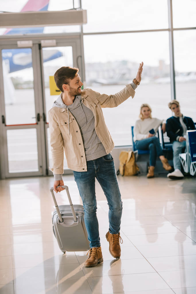 χαρούμενος νεαρός με βαλίτσα κουνώντας το χέρι και να ψάχνει μακριά στο αεροδρόμιο - Φωτογραφία, εικόνα