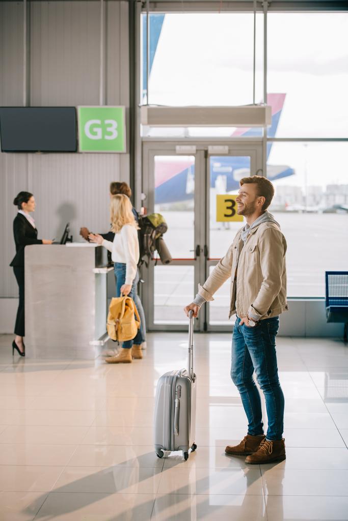 вид збоку усміхненого молодого чоловіка, що стоїть з валізою в аеропорту
 - Фото, зображення