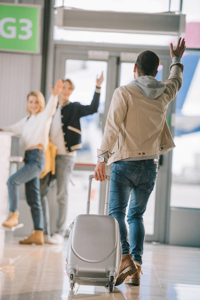 вид ззаду чоловіка з валізою махає рукою друзям на стійці реєстрації в аеропорту
 - Фото, зображення