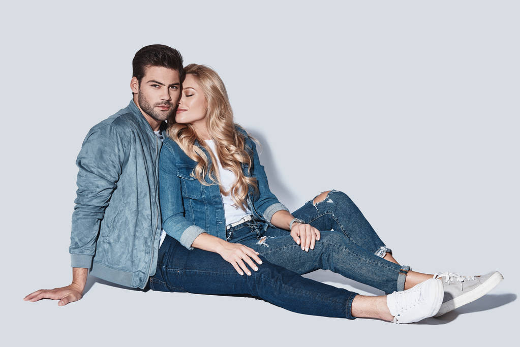 красивая молодая пара в джинсовой одежде сидит на полу на сером фоне
 - Фото, изображение