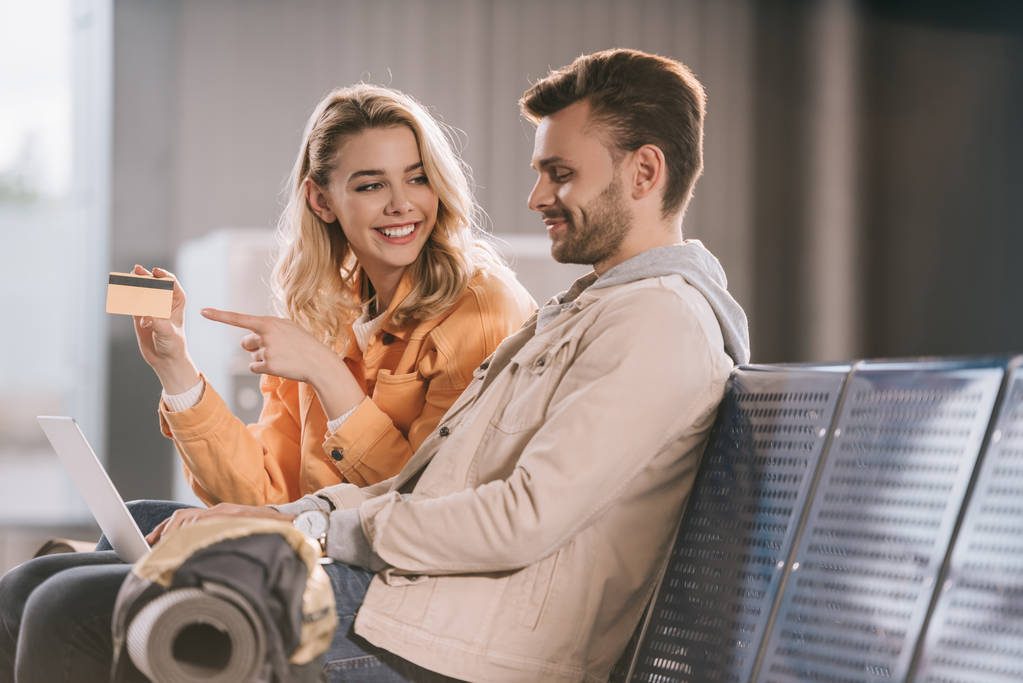 glimlachend jonge vrouw aan te wijzen met de vinger naar creditcard en op zoek naar man met laptop in luchthaven  - Foto, afbeelding