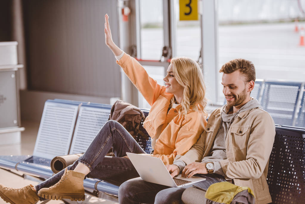 souriant jeune homme utilisant ordinateur portable et femme agitant la main tout en étant assis et en attendant ensemble à l'aéroport
 - Photo, image