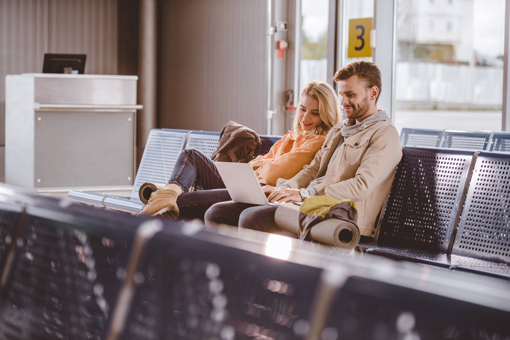 hymyilevä nuori pari käyttää kannettavaa tietokonetta ja odottaa lentoa lentokentällä
   - Valokuva, kuva