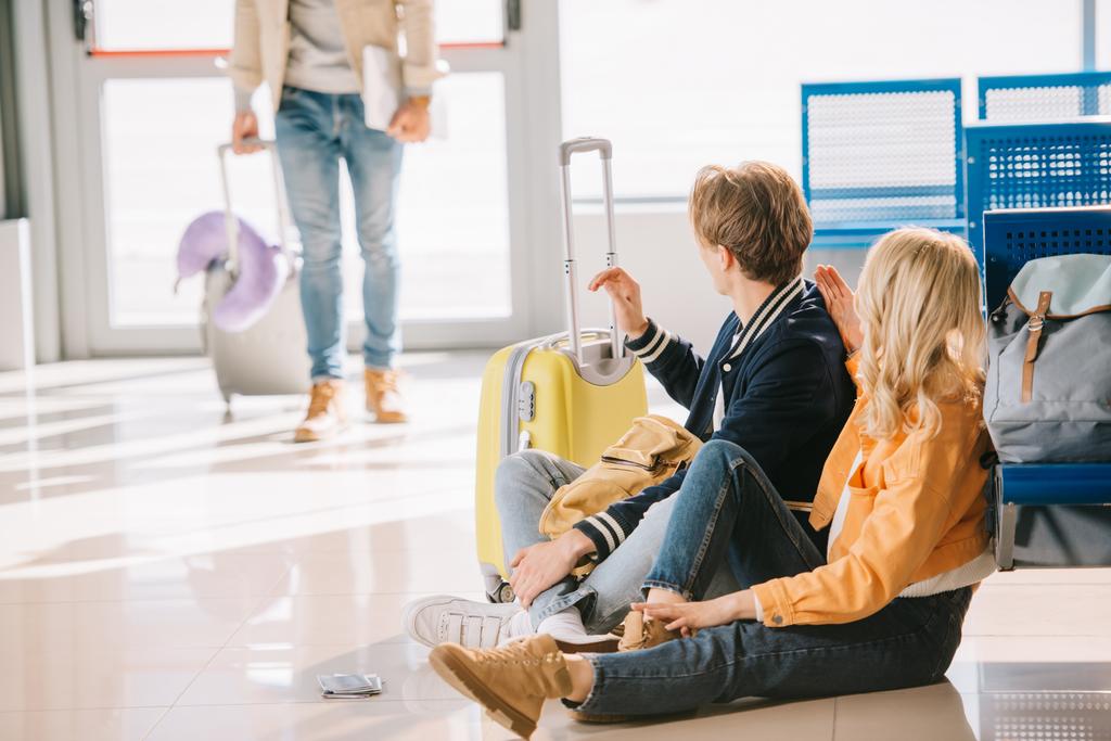 nuoret matkustajat istuvat lattialla ja katselevat ystäviä, jotka seisovat takana lentokentällä
  - Valokuva, kuva