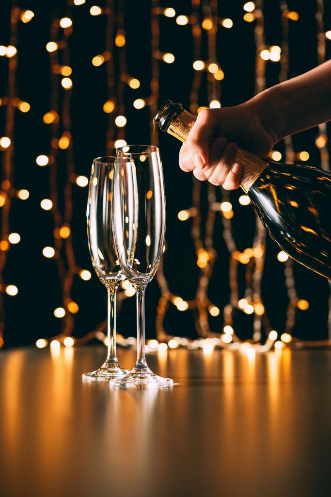 imagen recortada de la mujer sosteniendo botella de champán cerca de las copas en el fondo claro guirnalda, concepto de Navidad
 - Foto, Imagen
