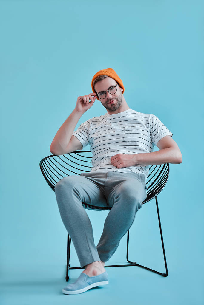 Knappe man geïsoleerd. Stijlvolle jongeman in brillen is poseren zittend op de stoel op blauwe achtergrond. - Foto, afbeelding