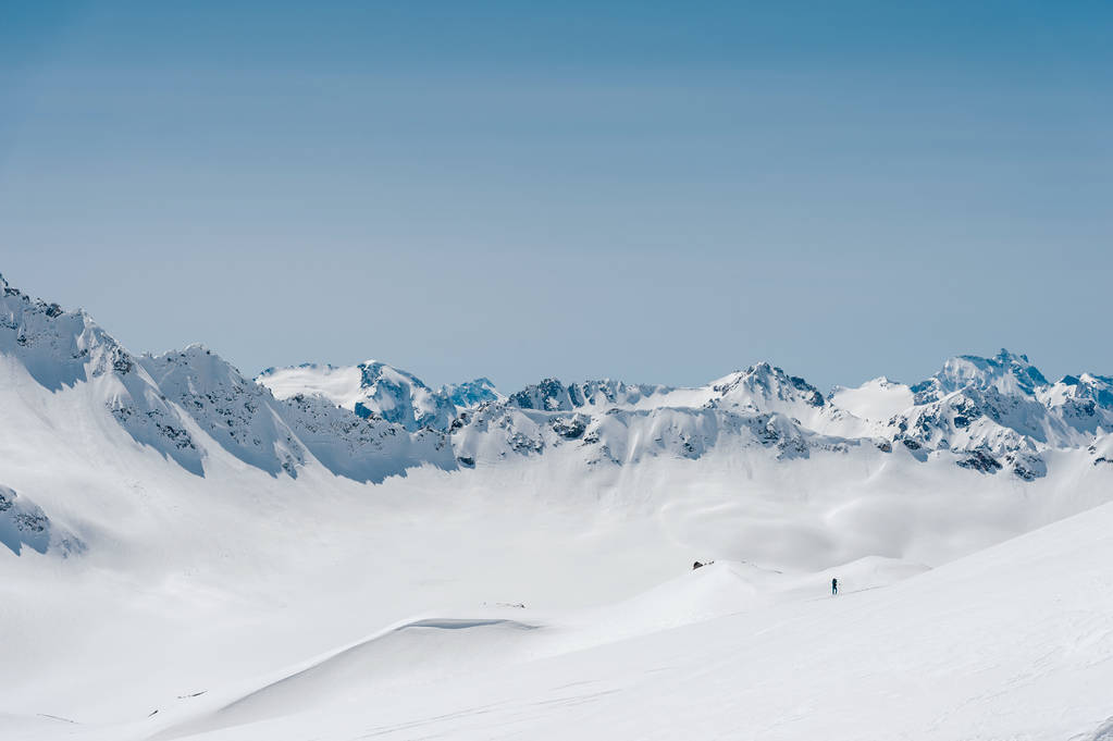 Ο ορειβάτης στέκεται στην άκρη του παγετώνα με ένα φτυάρι χιονιού στα χέρια του και δείχνει Shaks χειρονομία ενάντια σε το γαλάζιο του ουρανού και τα όρη του Καυκάσου - Φωτογραφία, εικόνα