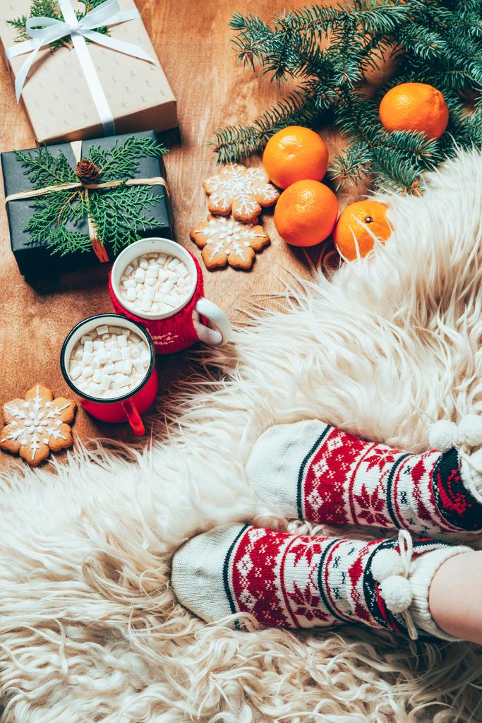 Teilansicht einer Frau in Socken im Hintergrund mit Tassen Kakao, Mandarinen, Plätzchen und Weihnachtsgeschenken - Foto, Bild