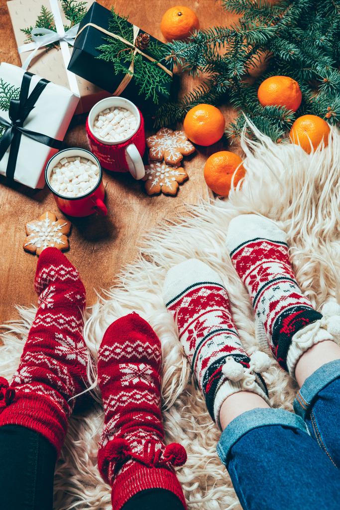 περικοπεί shot των γυναικών στο χειμώνα κάλτσες στο παρασκήνιο με τυλιγμένα δώρα Χριστουγέννων και φλιτζάνια ζεστή σοκολάτα με ζαχαρωτά - Φωτογραφία, εικόνα
