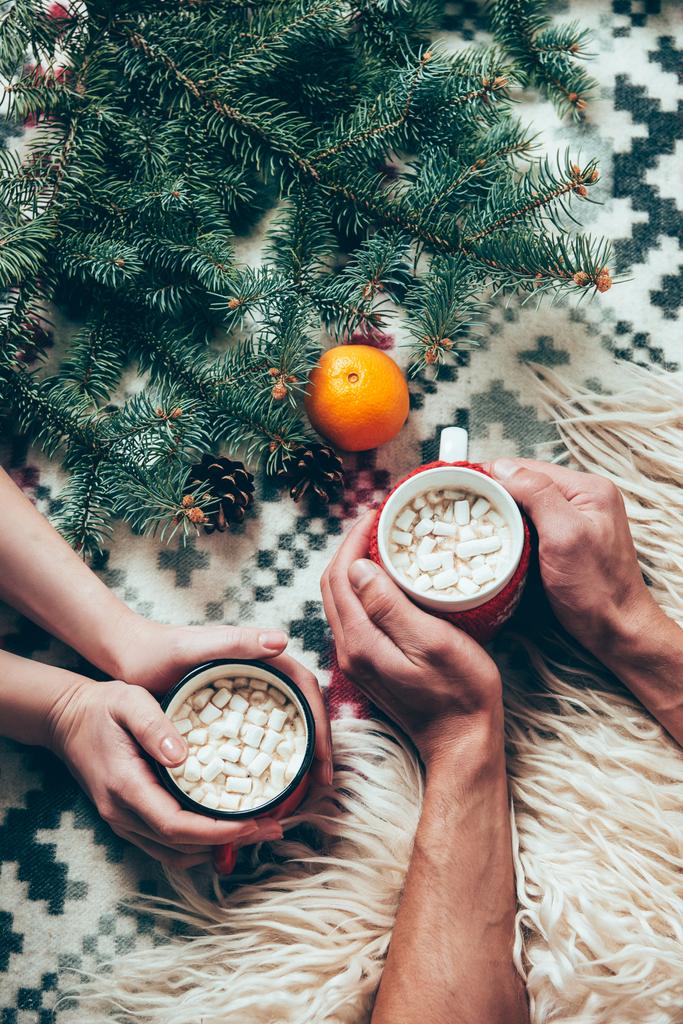colpo ritagliato di coppia che tiene tazze di bevande calde al cacao sullo sfondo con rami di pino e mandarino
  - Foto, immagini