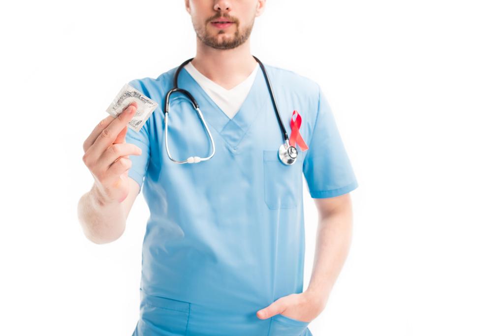 bijgesneden afbeelding van arts met een rood lint op uniforme weergegeven: condoom geïsoleerd op wit, wereld aids dag concept - Foto, afbeelding