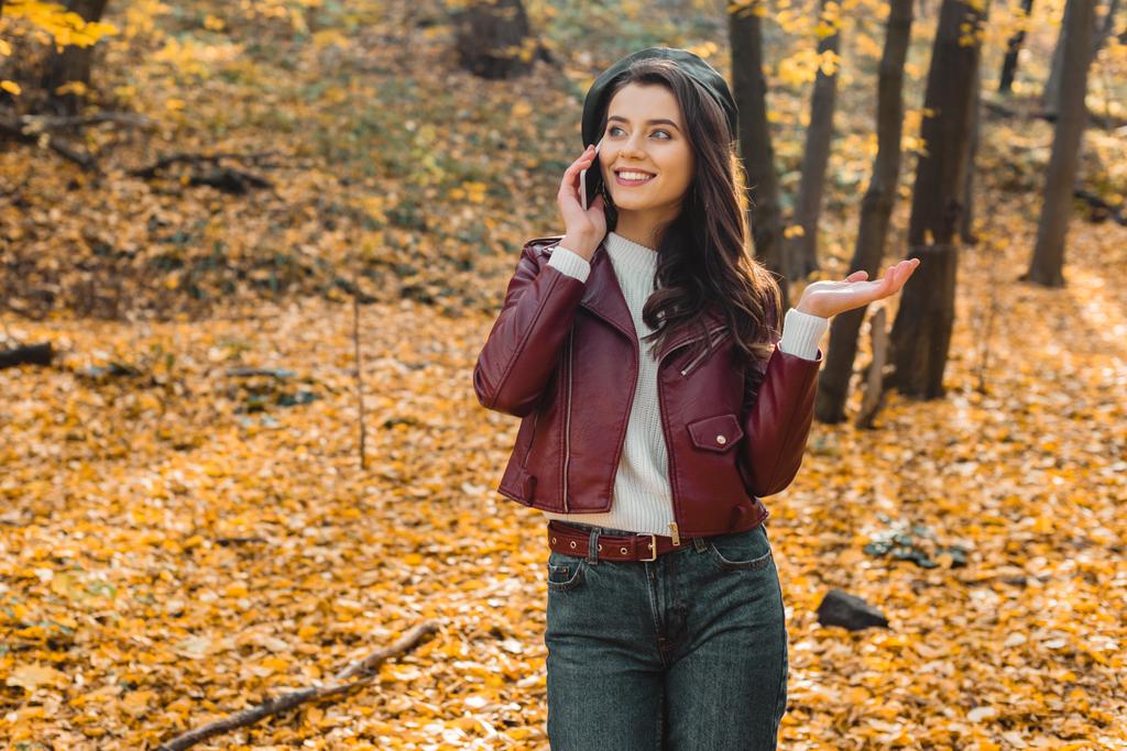 young stylish woman  in leather jacket talking on smartphone and gesturing by hand in autumnal park - Photo, Image