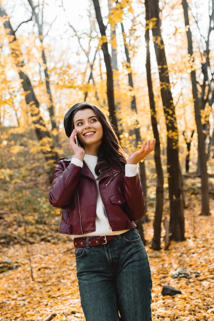 lage hoekmening van gelukkig meisje in lederen jas praten op smartphone en gebaren met de hand in de herfst park - Foto, afbeelding