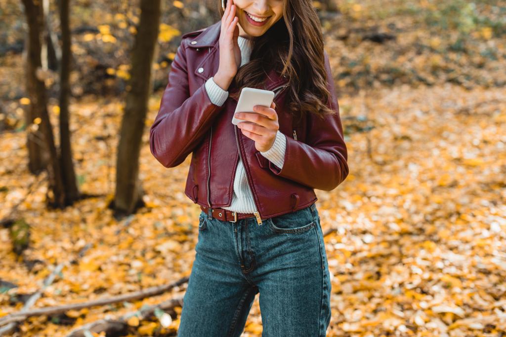 image recadrée de femme souriante dans une veste en cuir élégant en utilisant un smartphone dans la forêt automnale
 - Photo, image