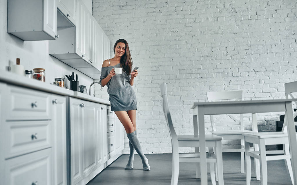 Goedemorgen! Aantrekkelijke jonge vrouw heeft rust thuis. Full-length beeld van meisje op keuken. - Foto, afbeelding