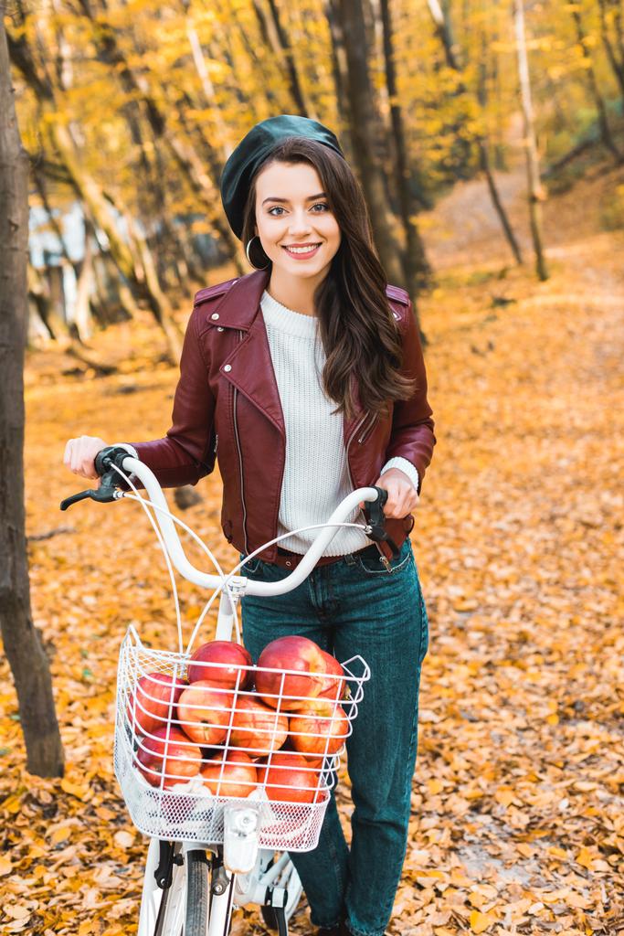 lächelndes stilvolles Mädchen in Lederjacke mit Fahrrad und Korb voller roter Äpfel im herbstlichen Park - Foto, Bild