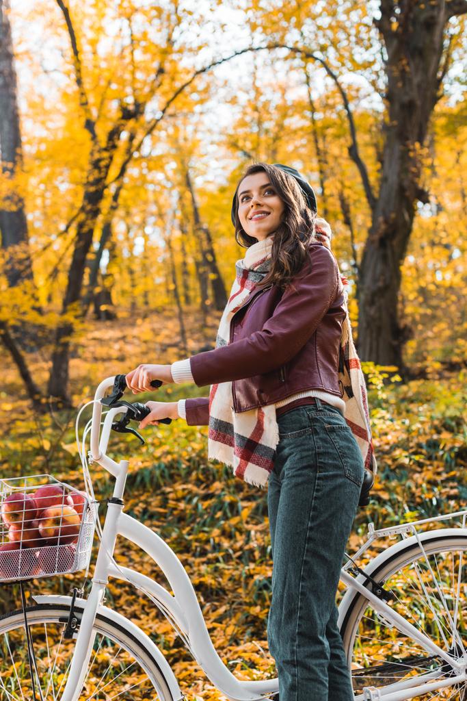 low angle view of smiling woman in stylish leather jacket and beret carrying bicycle in autumnal forest - Photo, Image