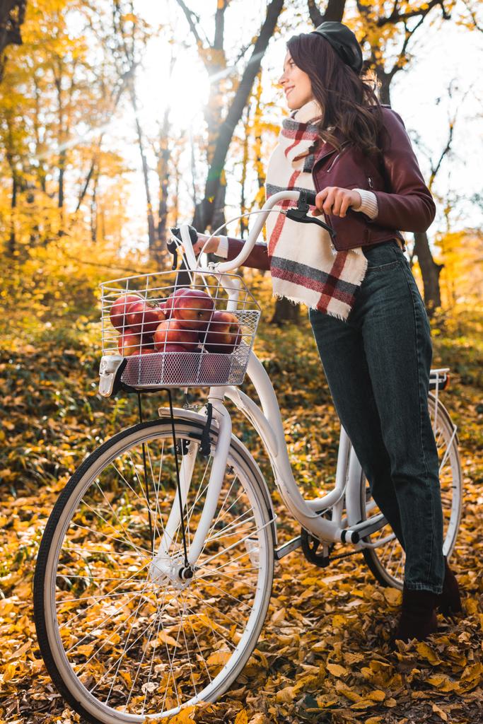 Tiefansicht einer stilvollen Frau in Lederjacke und Baskenmütze, die ein Fahrrad im gelben herbstlichen Wald trägt - Foto, Bild